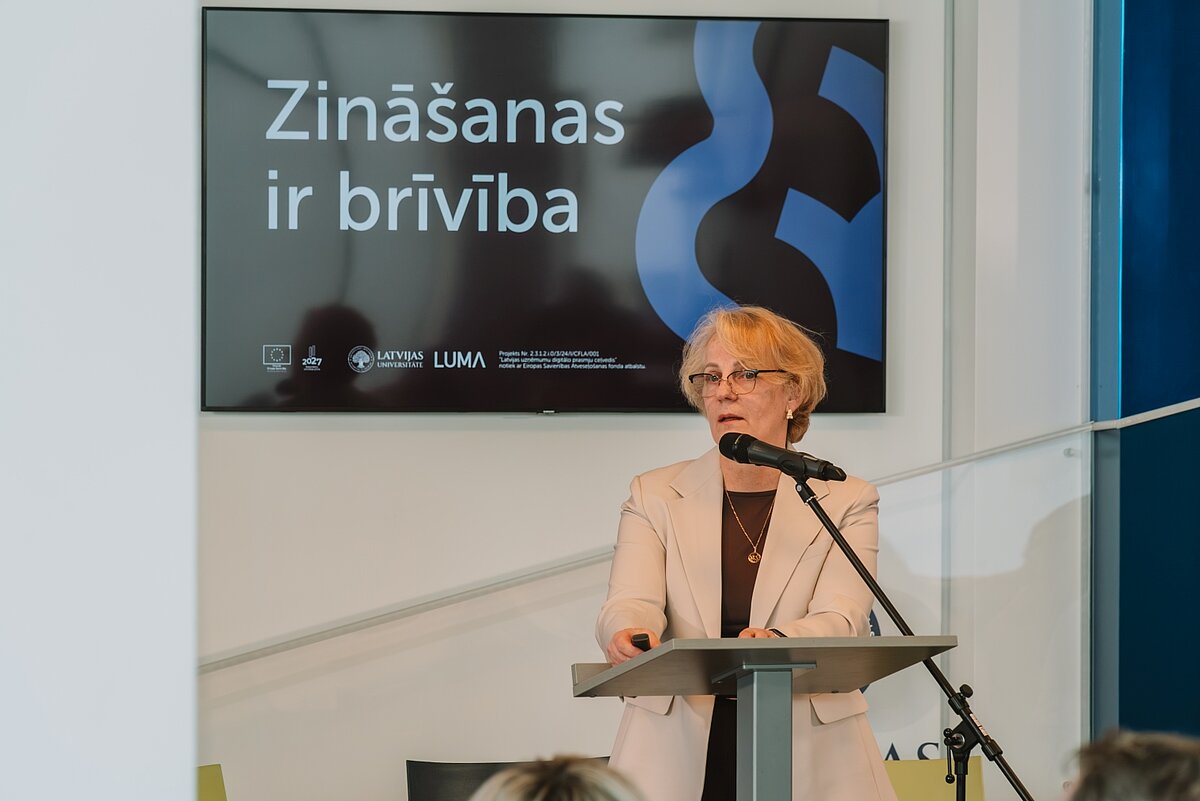 Sieviete ar brillēm bēšā žaketē uzstājas pie mikrofona. Fonā ekrāns ar "Zināšanas ir brīvība" un LU, LUMA logotipi.
