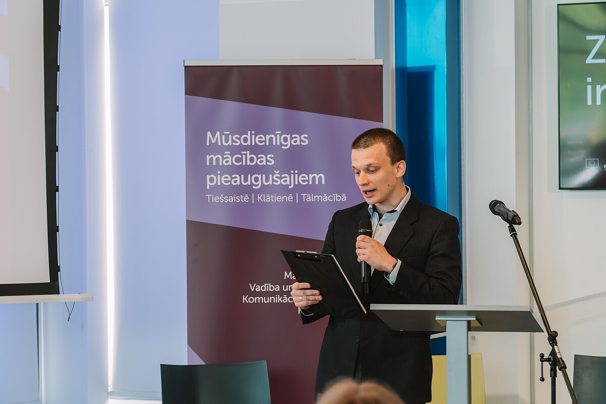 Moderators ar mikrofonu un mapi uzstājas LUDPC noslēguma pasākumā. Fonā LUMA roll-up baneris.
