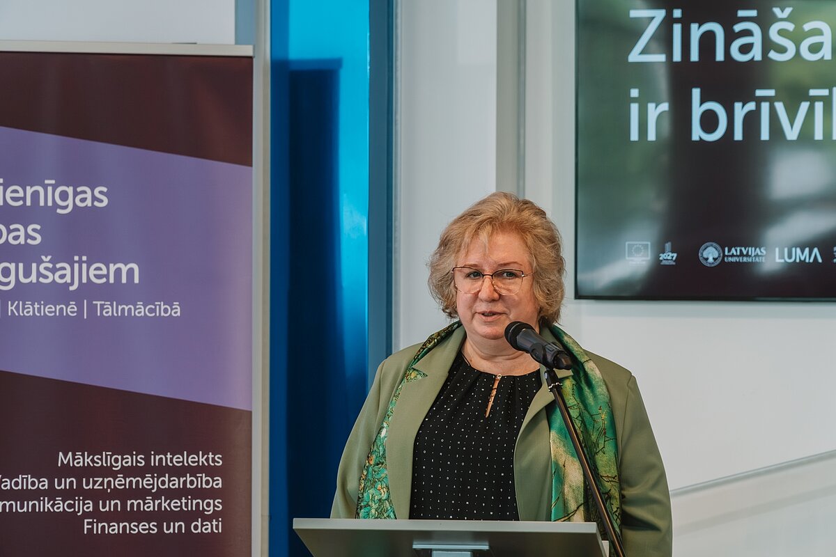 Sieviete ar brillēm un zaļu lakatiņu uzstājas pie mikrofona LUDPC noslēguma pasākumā. Fonā LUMA rullbaneris un "Zināšanas ir brīvība" ekrāns.